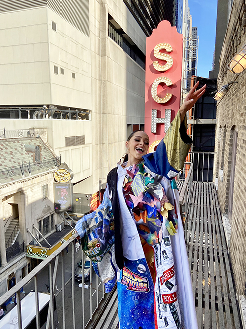Vazquez standing outside in daylight in front of the Schoenfeld Theatre marquis sign, smiling in the Robe with one arm up.