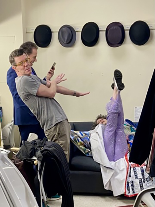 On the wall are a row of hats hanging up. Below it, Faure lies on a couch, her back on the seat, her arms outstretched and her legs straight up in the air. Two men look on amused, one of them (Gavin Lee), leaning away with his arms stretched towards her.